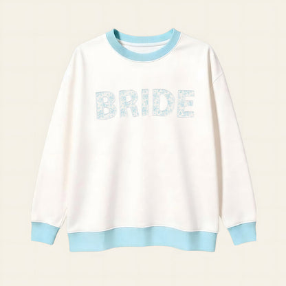The Darling Effect Bride Oversized Crew is a cozy white sweatshirt with a light blue collar, cuffs, and hem. "BRIDE" appears in large light blue floral letters on a soft cream grid background.