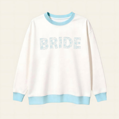 The Darling Effect Bride Oversized Crew is a cozy white sweatshirt with a light blue collar, cuffs, and hem. "BRIDE" appears in large light blue floral letters on a soft cream grid background.