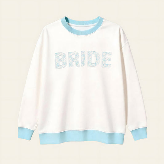 The Darling Effect Bride Oversized Crew is a cozy white sweatshirt with a light blue collar, cuffs, and hem. "BRIDE" appears in large light blue floral letters on a soft cream grid background.