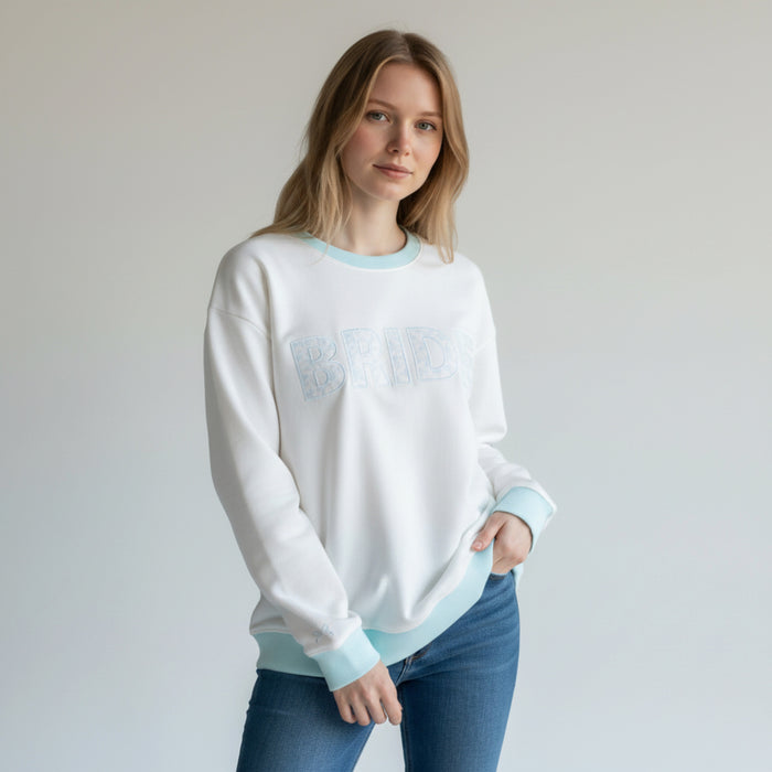 A woman with long blonde hair wears The Darling Effect's Bride Oversized Crew, a white sweatshirt with BRIDE embroidery, paired with blue jeans against a plain light background.