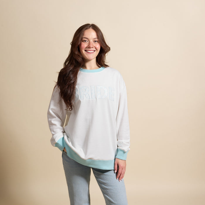 A smiling woman with long brown hair wears The Darling Effect’s Bride Oversized Crew, a cozy white sweatshirt with embroidered "BRIDE" lettering, paired with light blue jeans against a plain beige background.