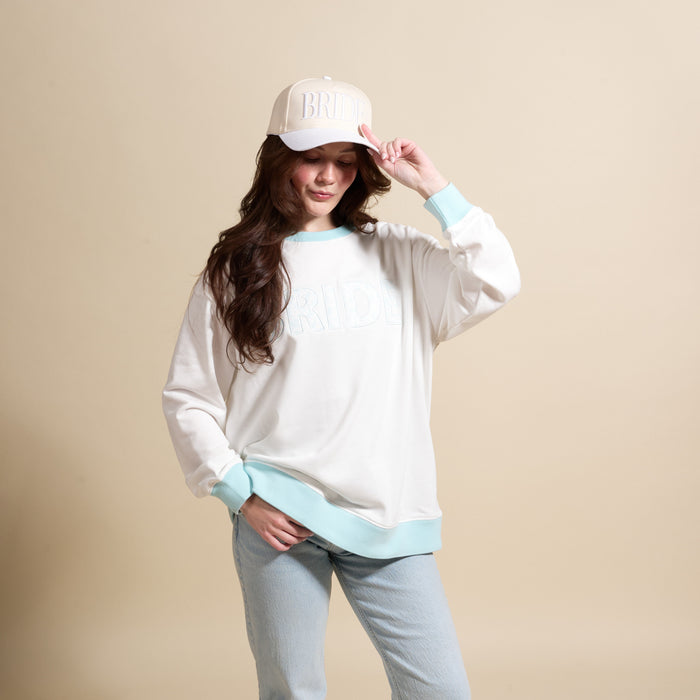 A woman in The Darling Effect’s Bride Oversized Crew and matching baseball cap poses against a beige background, smiling while holding her cap. She has long dark hair and is wearing light blue jeans.