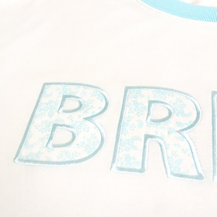 Close-up of light blue "BR" letters with a floral pattern, embroidered on The Darling Effect's Bride Oversized Crew. The soft white fabric and light blue trimmed collar make this sweatshirt cozy and stylish.