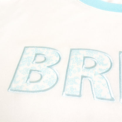 Close-up of light blue "BR" letters with a floral pattern, embroidered on The Darling Effect's Bride Oversized Crew. The soft white fabric and light blue trimmed collar make this sweatshirt cozy and stylish.
