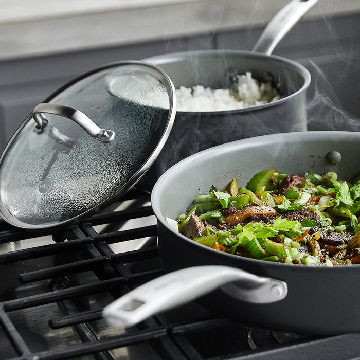 Gray ceramic 10-piece cookware set on a stovetop, with one pan holding vegetables and a pot containing rice.