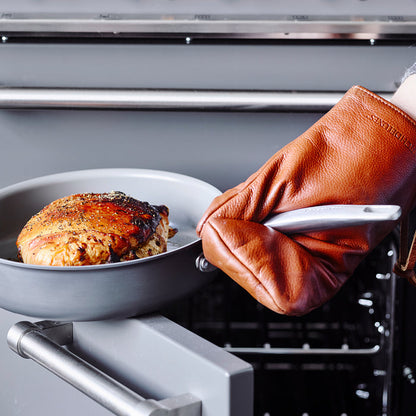 A brown oven mitt holds a skillet from the Chatham Ceramic Nonstick 10-Piece Set by an open oven.