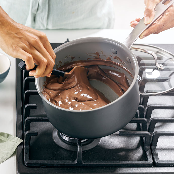 Melted chocolate being stirred in a gray ceramic nonstick saucepan with lid on a stove.