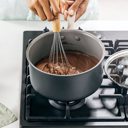 Chocolate pudding being whisked in a ceramic nonstick saucepan with lid on stove, cloth beside the pan.