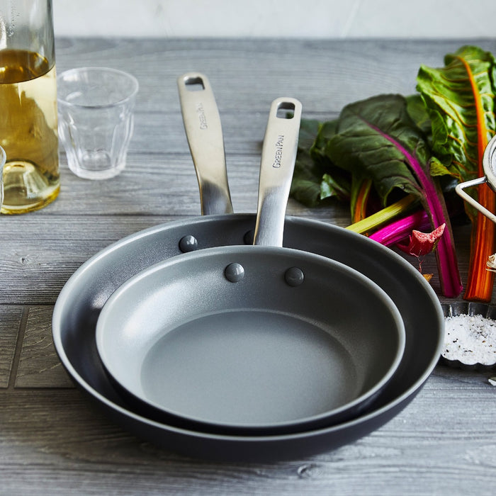 Two stacked ceramic nonstick frypans, 8 and 10, on wood beside leafy greens and a bottle of oil.