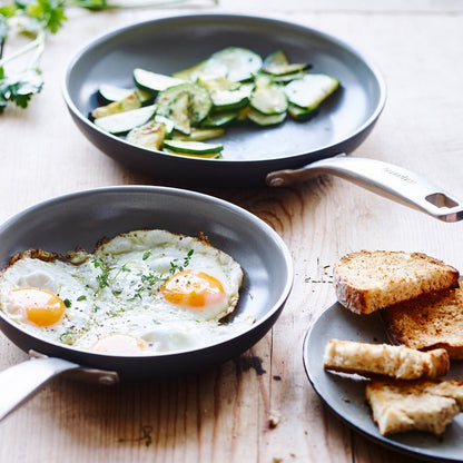 Two ceramic nonstick frypans, 8 and 10, cooking eggs and zucchini, next to toast on a wooden table.