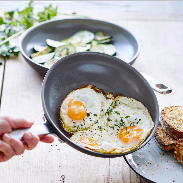 Hand holds a ceramic nonstick frypan with three sunny-side-up eggs; vegetables and toast sit beside the pan.