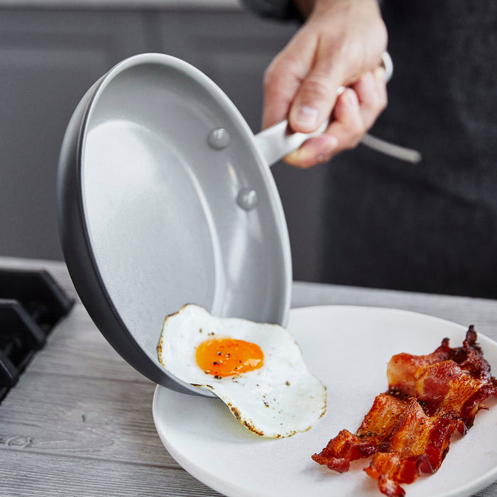 A hand slides a fried egg from a ceramic nonstick frypan onto a plate; bacon is on the counter nearby.