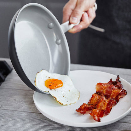 A hand slides a fried egg from an 8-inch ceramic nonstick frypan onto a plate with bacon strips.