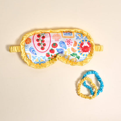 The Darling Effect’s Ciao Bella Beauty Sleep Mask features coastal prints, yellow ruffle trim, and comes with two small satin scrunchies in bright yellow and blue.