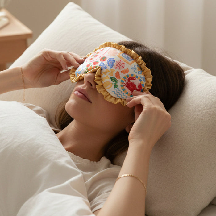 A woman in a softly lit room adjusts The Darling Effect’s Ciao Bella Beauty Sleep Mask, featuring ruffled edges, as she rests her head on a white pillow.