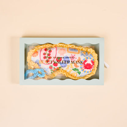 The Darling Effect Ciao Bella Beauty Sleep Mask set features a floral satin mask with yellow trim, blue satin scrunchie, and comes in a light green box with clear window. "Beauty Sleep Mask" is printed on the mask.