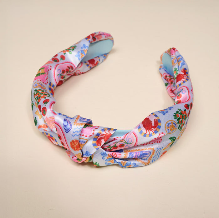 The Darling Effect’s Ciao Bella Classic Twist Headband features shiny, satin-like fabric with a vibrant fruit and motif pattern on light blue and pink, making it a versatile accessory displayed on a beige surface.