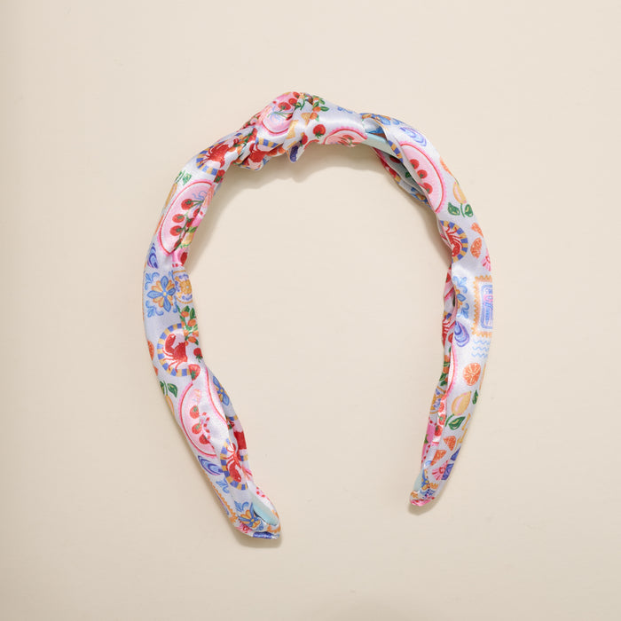The Darling Effect’s Ciao Bella Classic Twist Headband features a knotted top and vibrant floral-paisley print in red, blue, green, and yellow on white—a stylish, versatile accessory displayed on a beige surface.