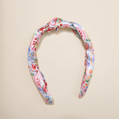 The Darling Effect’s Ciao Bella Classic Twist Headband features a knotted top and vibrant floral-paisley print in red, blue, green, and yellow on white—a stylish, versatile accessory displayed on a beige surface.