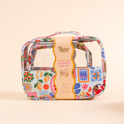The Darling Effect's Ciao Bella Clearly Chic Cosmetic Bag Set features clear bags with colorful floral and fruit designs, packaged with a tan label. This portable toiletry duo is shown on a light beige background.