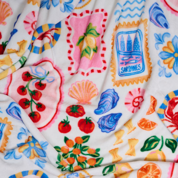 The Darling Effect’s Ciao Bella Snuggle Up Plush Blanket features bright illustrations of tomatoes, oranges, sardines, lemons, seashells, and flowers in vibrant red, blue, yellow, orange, and pink on a soft white background.