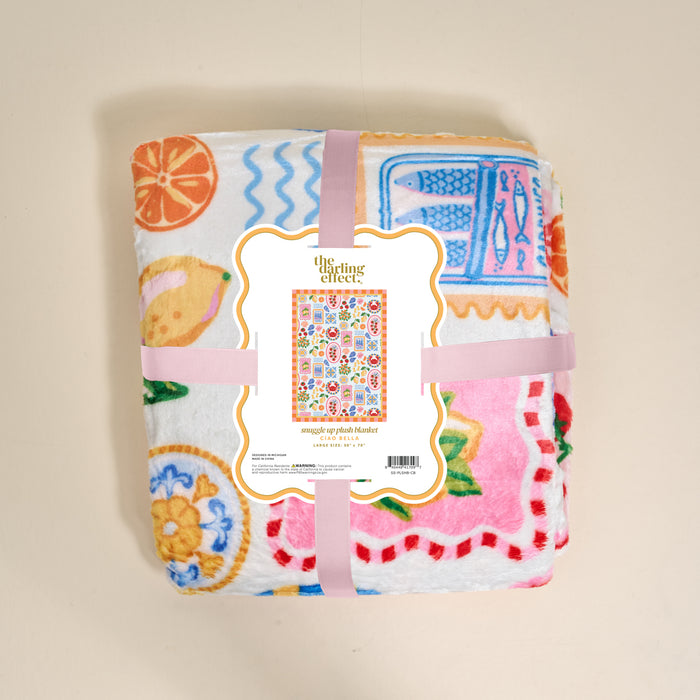 A folded, colorful Ciao Bella Snuggle Up Plush Blanket by The Darling Effect features fruit and kitchenware prints, wrapped in a pink ribbon. The label displays the brand name and a small image of the blanket’s full design.