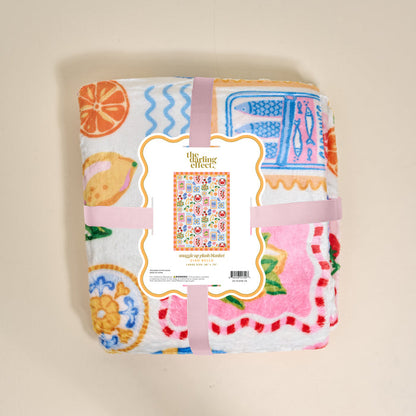 A folded, colorful Ciao Bella Snuggle Up Plush Blanket by The Darling Effect features fruit and kitchenware prints, wrapped in a pink ribbon. The label displays the brand name and a small image of the blanket’s full design.