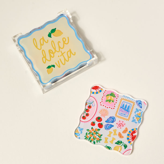 The Darling Effect’s Ciao Bella Table Talk Acrylic Coaster Set features two decorative scalloped-edge coasters—one in a clear holder with “la dolce vita” and lemons, one with colorful Italian-inspired art—perfect for any table.