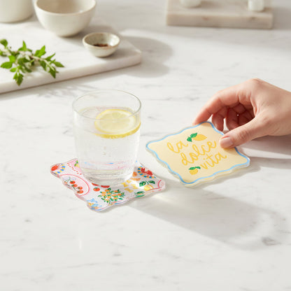 A hand picks up a Ciao Bella Table Talk Acrylic Coaster by The Darling Effect, featuring "la dolce vita," next to a lemon sparkling water on another coaster, set on marble with herbs and bowls for charming table décor.