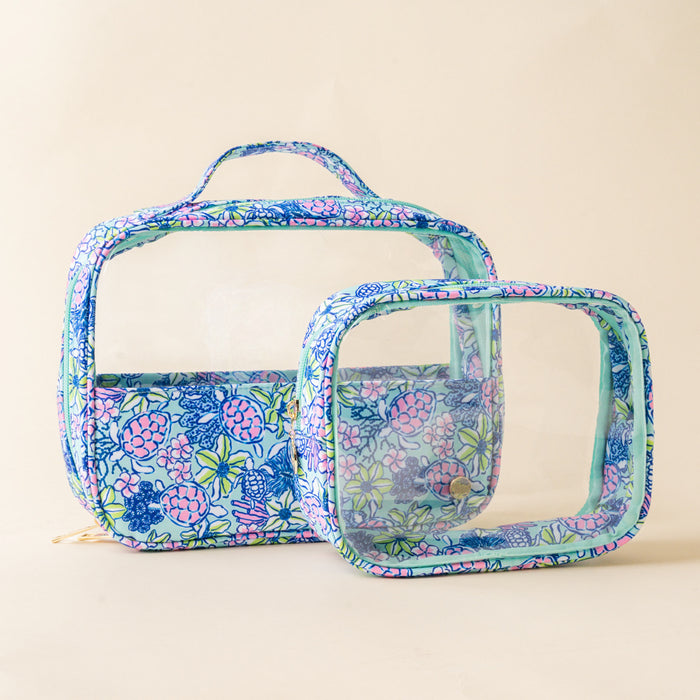 Two clear makeup bags trimmed in a blue, pink, and green floral sea turtle print. This cosmetic bag set includes a larger handled train case and a smaller square pouch, both featuring transparent panels and gold zipper hardware.