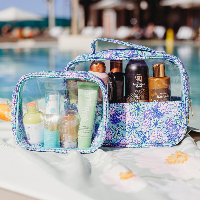 The Coastal Cove Clearly Chic Cosmetic Bag Set by the-darling-effect, featuring one clear and one opaque floral-patterned bag, stylishly holds sunscreen, skincare, and hair products poolside with a sunhat in view.