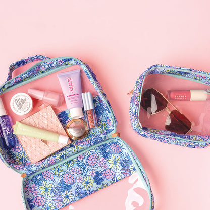 The-darling-effect’s Coastal Cove Clearly Chic Cosmetic Bag Set features two floral-patterned bags on a pink backdrop—one open with skincare and makeup, the other holding sunglasses and a lip gloss.