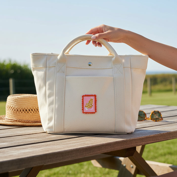 A hand holds the handle of a white canvas tote bag featuring The Darling Effect’s Conch Mix & Match Iron-On Patch, resting on a wooden picnic table outdoors beside a straw hat and sunglasses under sunny skies.