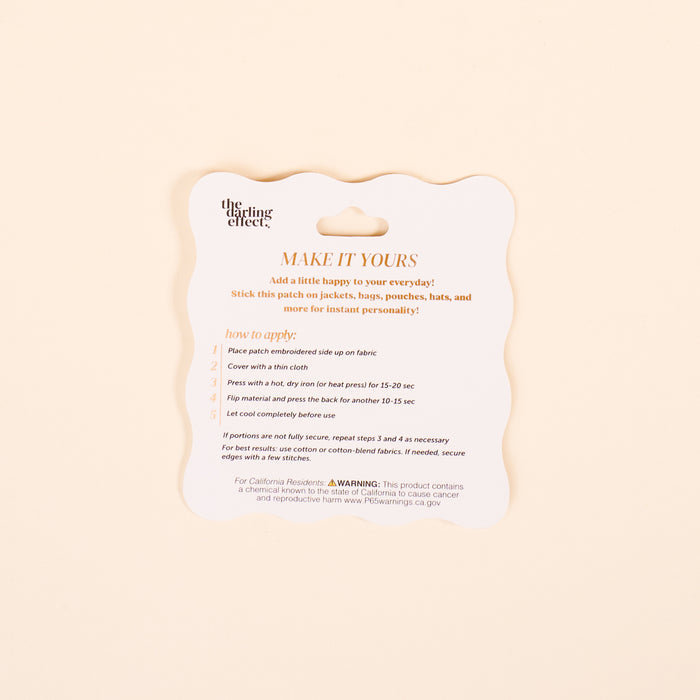 A square scalloped-edge card titled MAKE IT YOURS from The Darling Effect explains how to use the Conch Mix & Match Iron-On Patch, with steps and safety tips on a light beige background—ideal for customizing jackets with colorful designs.