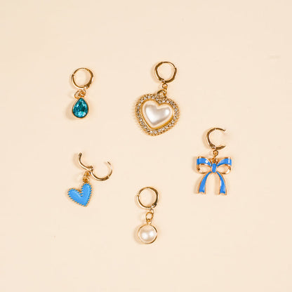 The Darling Effect’s Coquette Cloud The Ultimate Charm Pack features five gold hoop earrings with unique charms—blue teardrop, pearl heart with rhinestones, stitched blue heart, round pearl, and blue ribbon—perfect for mixing and matching.