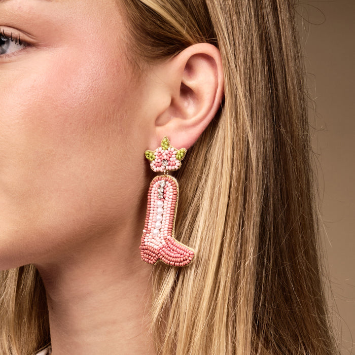 A woman with straight blonde hair wears The Darling Effect’s Cowgirl Boot Beaded Earrings—vibrant pink, hand-beaded with a floral detail. Her face is partially visible against a neutral background.
