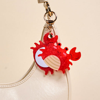 The Darling Effect's Crab Mini Mirror Bag Charm, shaped like a cute red crab, hangs from the handle of a white handbag against a plain beige background.