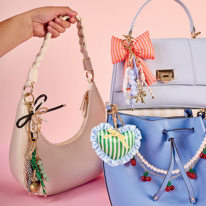 Three handbags—cream, pale purple, and blue—from The Darling Effect are decorated with bead, bow, pearl, striped heart, cherry accents, and the Crab Mini Mirror Bag Charm. Displayed on a pink background; a hand holds the cream bag.