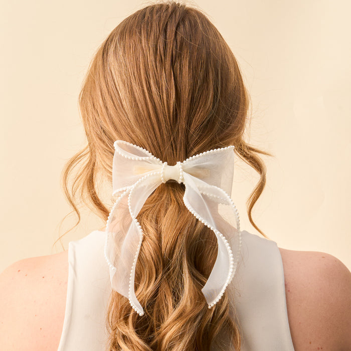 A person with long, wavy, light brown hair is shown from behind wearing the-darling-effect's Cream Sheer Pearl Hair Bow, a delicate clip with decorative edges. They pair it with a sleeveless white top, highlighting this elegant accessory.
