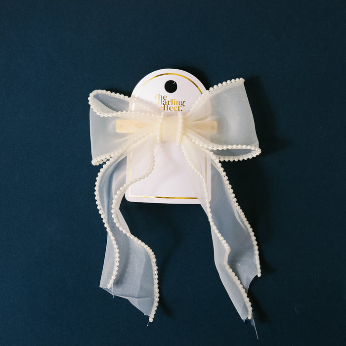 The Cream Sheer Pearl Hair Bow by the-darling-effect features an ivory sheer design with pearl trim, attached to a white card with the brand’s logo—a charming addition to your hair accessories.