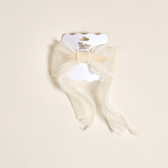 The Cream Sheer Pearl Hair Bow by the-darling-effect features a delicate cream bow with beaded trim, attached to a white branded card and displayed on a light beige background—ideal for hair accessory enthusiasts.