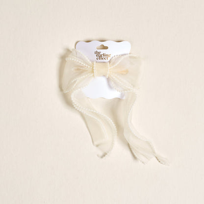 The Cream Sheer Pearl Hair Bow by the-darling-effect features a delicate cream bow with beaded trim, attached to a white branded card and displayed on a light beige background—ideal for hair accessory enthusiasts.