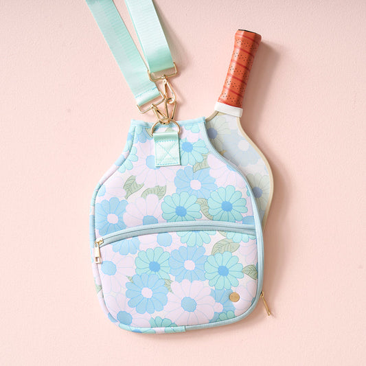 The Darling Effect's Daisy Craze Aqua Pickleball Paddle Case, featuring blue and green floral patterns, hangs on a pink wall. A paddle with a tan handle is partially inside the case, which includes a front zip pocket and adjustable strap.