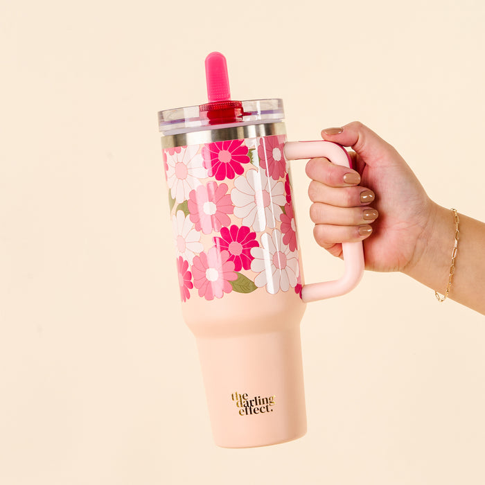 A hand holds the-darling-effect Daisy Craze Hot Pink 40 oz Lifestyle Flip Straw Tumbler with a retro floral pattern, pink handle, leak-resistant clear lid, and pink straw. “the darling effect.” logo is on the tumbler bottom.