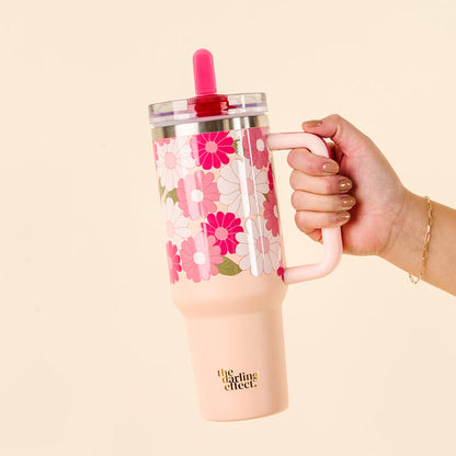 A hand holds the-darling-effect Daisy Craze Hot Pink 40 oz Lifestyle Flip Straw Tumbler with a retro floral pattern, pink handle, leak-resistant clear lid, and pink straw. “the darling effect.” logo is on the tumbler bottom.