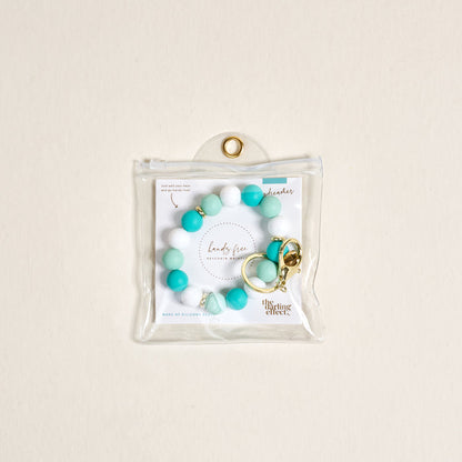 The Darling Effect’s Daydreamer Hands-Free Keychain Wristlet features teal and white silicone beads with a gold keyring, packaged in a clear pouch against a light beige background—stylish convenience for on-the-go use.