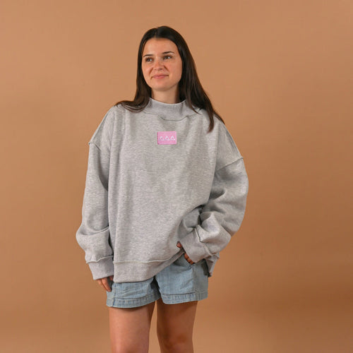 A woman with long brown hair stands against a tan background wearing The Darling Effect's Delta Delta Delta Letter Patch Mockneck Sweatshirt in L/XL and light denim shorts, hands in her pockets and a relaxed look on her face.