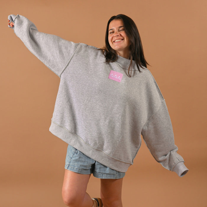 A smiling woman wears The Darling Effect's Delta Delta Delta Letter Patch Mockneck Sweatshirt (L/XL) with denim shorts, arms outstretched against a plain tan background.