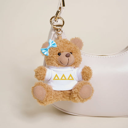 A small brown Delta Delta Delta Teddy Bear Bag Charm by The Darling Effect, with a blue bow and white shirt featuring three yellow triangles, hangs from a white handbag.