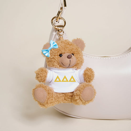 A small brown Delta Delta Delta Teddy Bear Bag Charm by The Darling Effect, with a blue bow and white shirt featuring three yellow triangles, hangs from a white handbag.
